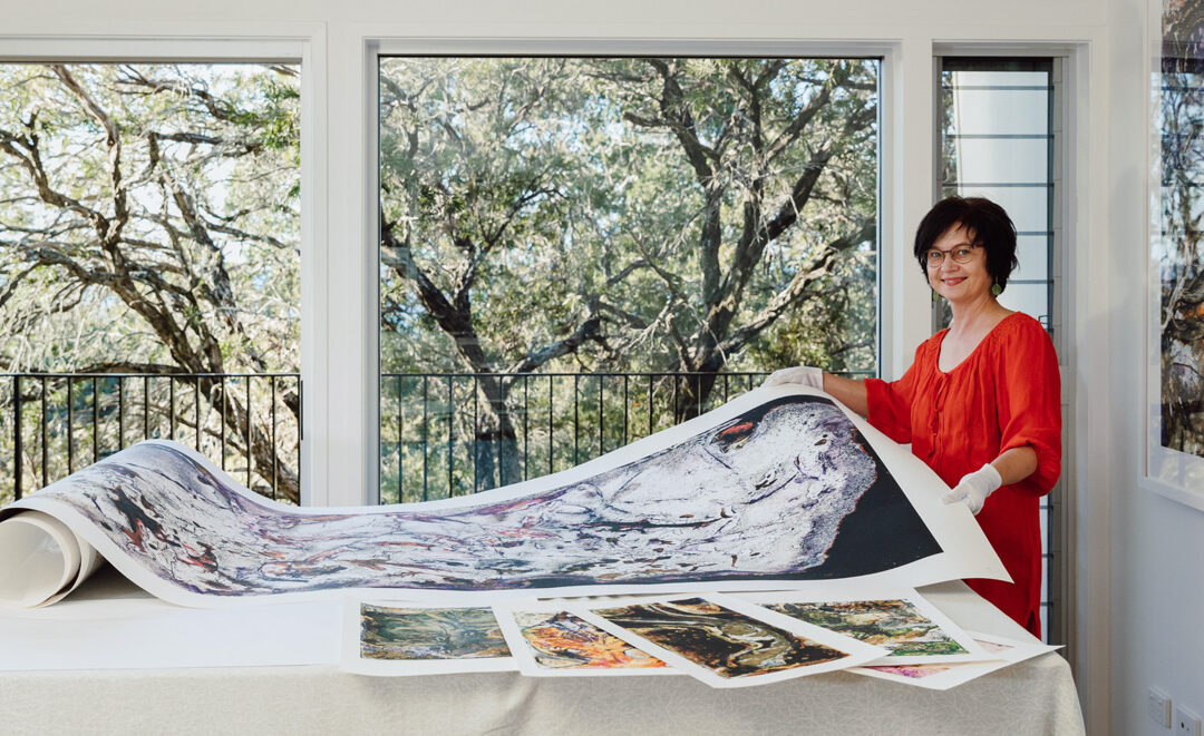 a dark hair woman in orange top staning in front of a large window shows a large print on paper laid out on a table